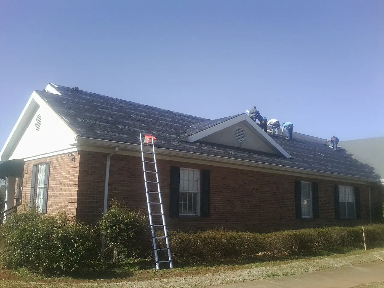Crew tearing shingles off a brick ranch roof with a ladder set — Metal Roof Repair work by our crew in Ackley