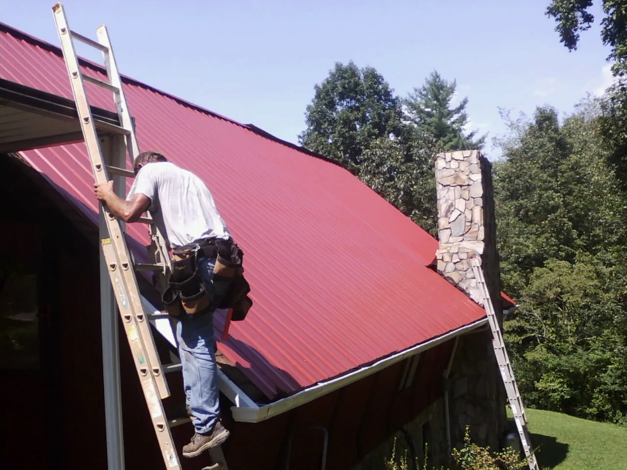 Roofer on a ladder beside a red metal roof and stone chimney — Soffit & Fascia Repair by Roofing Brothers in Ackley